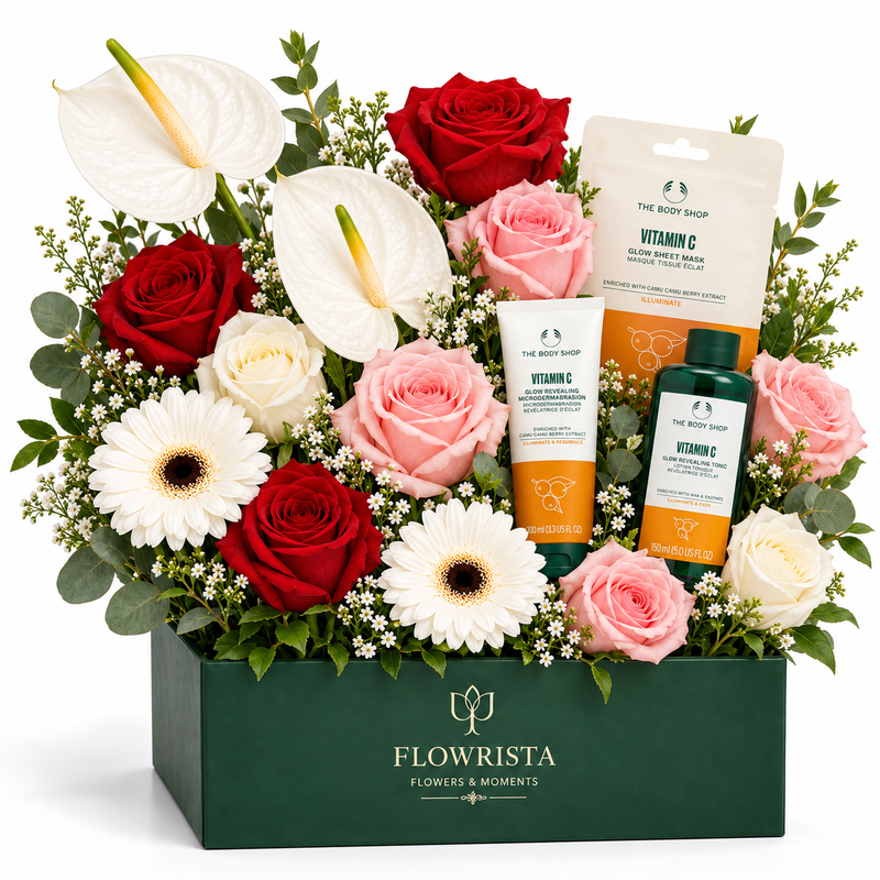 A floral gift box arrangement from Flowrista featuring red and pink roses, white and pink anthuriums, daisies, baby's breath, and three The Body Shop Vitamin C skincare products arranged in a dark green box.