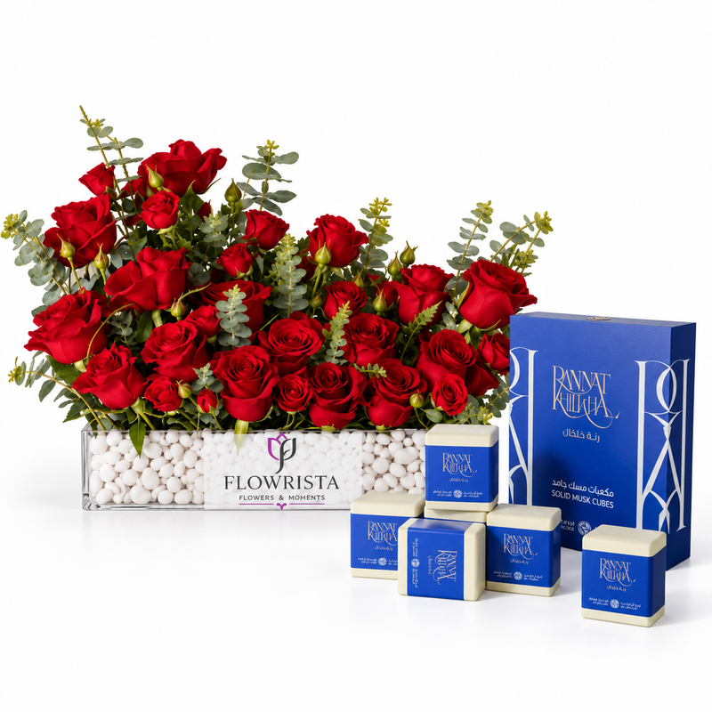 A floral gift arrangement featuring a large bouquet of red roses in a clear rectangular planter filled with white pebbles, alongside several blue and white boxed gift items branded RAINY RAINA. The arrangement is photographed on a clean white studio background.