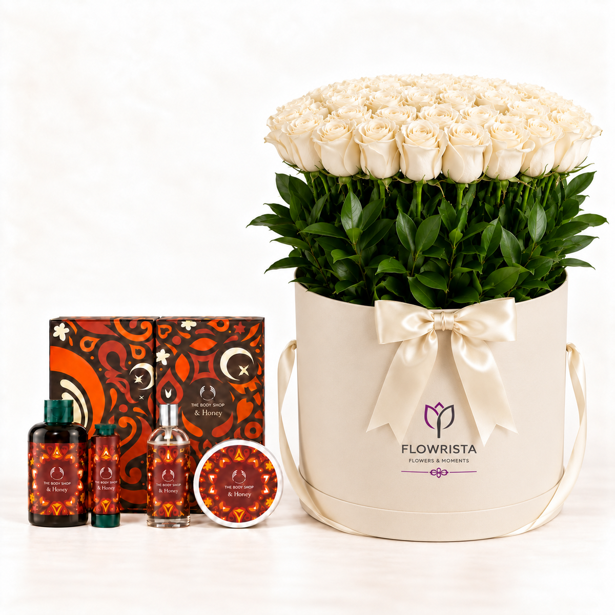 A luxury gift set featuring a large bouquet of 60 white roses in a cream-colored cylindrical box with a satin bow, alongside three small bottles and a tin of rose essential oil from the brand FLOWRISTA. The set is photographed on a clean white background.