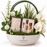 A curated gift basket arrangement by Flowwrista featuring small cosmetic bottles in a dark box, surrounded by white calla lilies, green foliage, and moss in a white ceramic basket with the Flowwrista logo.