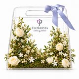 A clear acrylic flower gift bag with a lavender satin handle ribbon, filled with white roses, baby's breath, and green foliage. The bag features the 'FLOWRISTA FLOWERS & MOMENTS' logo printed on the front, set against a clean white background.
