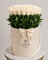 A large round cream-colored gift box filled with a dense arrangement of white roses and green stems, tied with a cream satin bow. The box is branded 'FLOWRISTA' and sits on a marble-topped table against a light gray background.