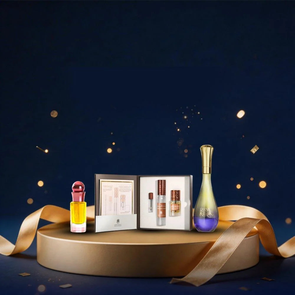 A luxury fragrance gift set displayed on a round gold pedestal against a dark navy blue background with gold bokeh lights and ribbon accents. The set includes a small yellow perfume bottle with a red cap, an open silver box containing three small cylindrical bottles, and a tall iridescent bottle with a gold cap and purple base.