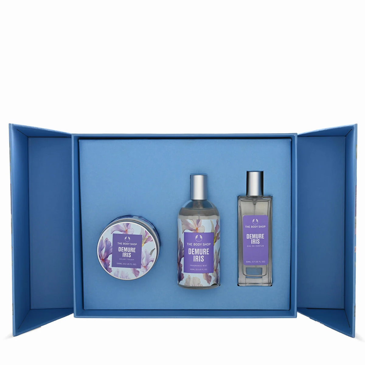 A perfume gift set from The Perfume Shop featuring three items: a round tin, a glass bottle, and a rectangular bottle, all labeled 'DEMIURE IRIS' with purple floral labels and silver caps, presented inside an open blue gift box on a white background.
