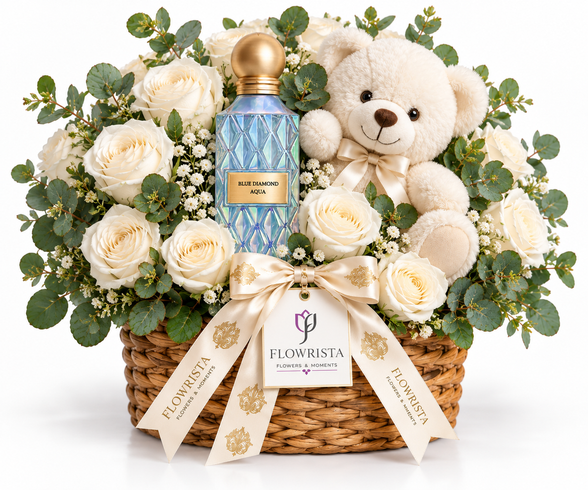 A luxury flower and gift basket arrangement featuring a blue patterned perfume bottle, a cream teddy bear, white roses, eucalyptus leaves, and baby's breath in a wicker basket, tied with a Flowrista ribbon.