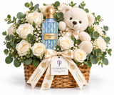 A luxury flower and gift basket arrangement featuring a blue patterned perfume bottle, a cream teddy bear, white roses, eucalyptus leaves, and baby's breath in a wicker basket, tied with a Flowrista ribbon.
