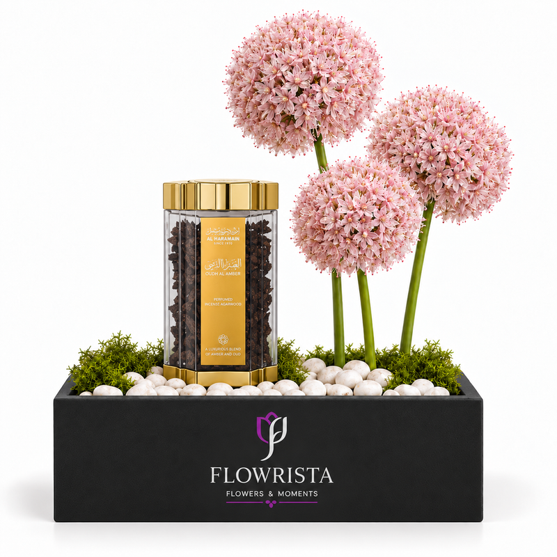 A floral gift arrangement in a black rectangular box from Flowrista, featuring three pink allium-like flower balls on green stems, white pebbles, green moss, and a clear cylindrical jar with gold trim containing dark tea leaves and a gold label.