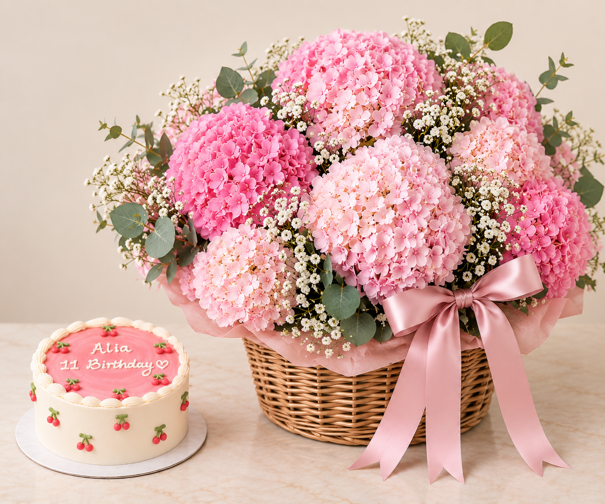 A large floral bouquet of pink hydrangeas and baby's breath in a wicker basket with a pink ribbon, displayed beside a small round birthday cake on a beige tabletop against a soft beige background.