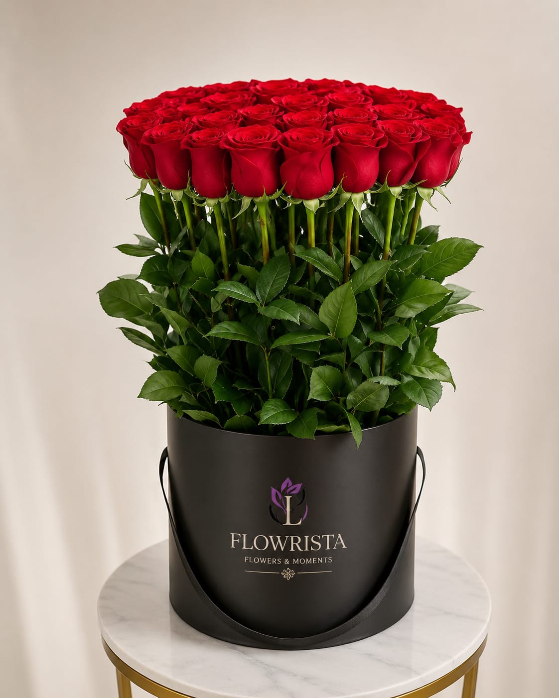A large bouquet of 60 fresh red roses with green stems and leaves, presented in a black cylindrical container with the brand name 'FLOWRISTA' printed on the front, set on a marble-topped table against a neutral beige background.