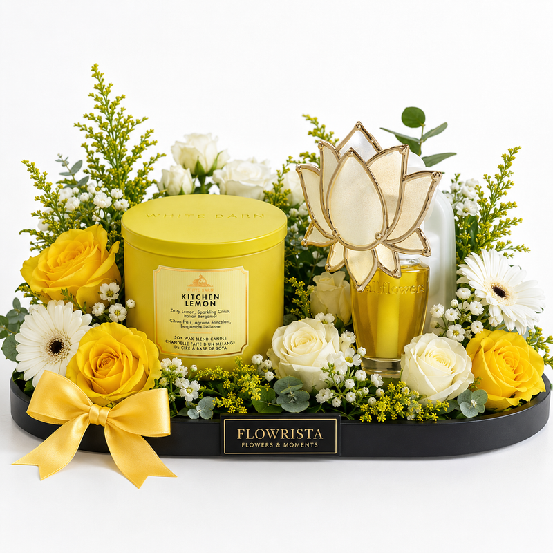 A curated floral gift arrangement on a black tray from Flowrista, featuring a yellow Kitchen Lemon scented candle tin, a gold-and-white lotus-shaped glass diffuser, yellow and white roses, daisies, green foliage, and a yellow ribbon bow, photographed on a white background.
