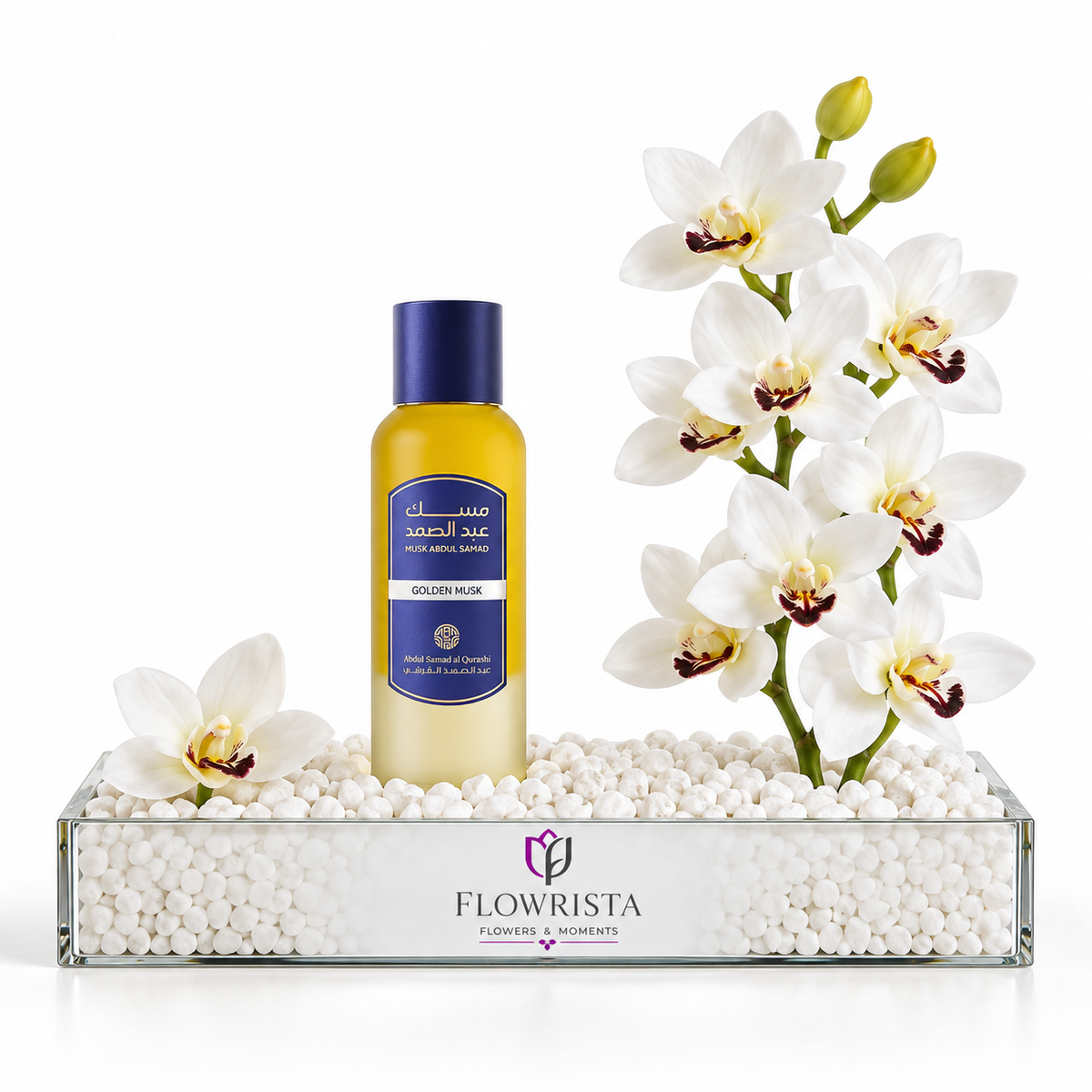 A bottle of Flowrista Golden Rose body wash standing in a clear glass tray filled with white decorative stones, styled with white orchids on a clean white background.