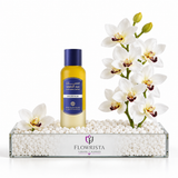 A bottle of Flowrista Golden Rose body wash standing in a clear glass tray filled with white decorative stones, styled with white orchids on a clean white background.