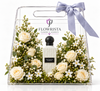 A clear acrylic gift box with a handle, tied with a lavender ribbon, containing a bottle of Flowrista 'White & Bright Vanilla' perfume surrounded by cream roses, white flowers, and green foliage on a white background.