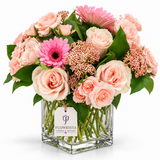 A vibrant floral arrangement in a square clear glass vase, featuring blush pink and cream roses, bright pink gerbera daisies, and green foliage, with a 'FLOWRISTA' tag on the vase, set against a solid white background.