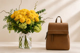 A brown suede-look backpack stands beside a glass vase filled with a bright yellow and green floral arrangement on a light beige surface against a cream wall with soft diagonal shadows.