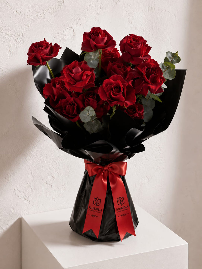 A bouquet of 12 red roses wrapped in black paper with a red ribbon bow, displayed on a white pedestal against a textured white wall.