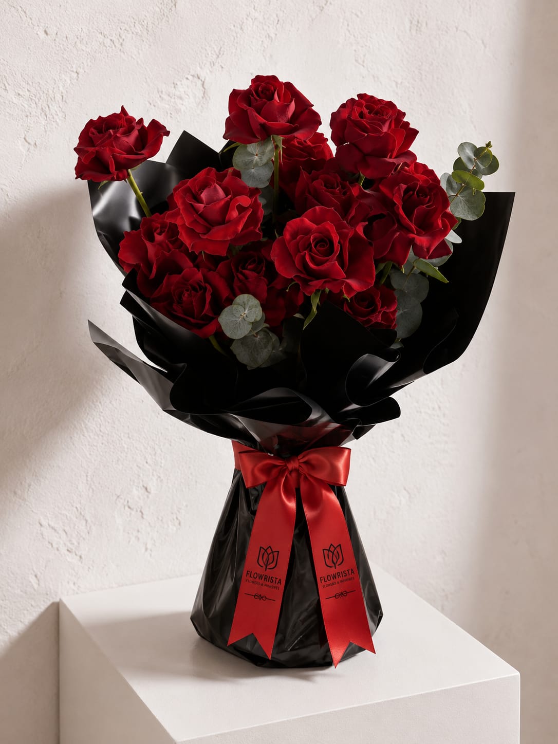 A bouquet of 12 red roses wrapped in black paper with a red ribbon bow, displayed on a white pedestal against a textured white wall.