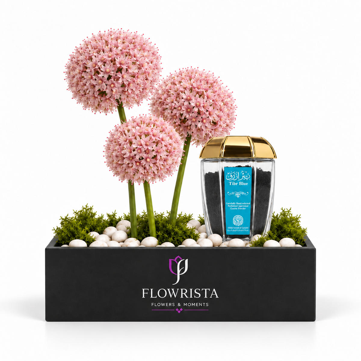 A flower arrangement gift set from Flowrista featuring three pink allium-like flower balls in a black rectangular planter with white stones and green moss, alongside a clear jar of Tile Blue potting mix with a gold lid, on a white background.
