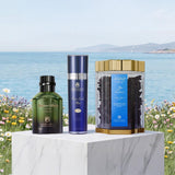 Three premium cosmetic products from the brand Alhambra are displayed on a white marble pedestal against a scenic background of the sea and flowers. The products include a dark green bottle of Amber Amber body oil, a blue bottle of Khamel Mist, and a clear jar with a gold lid containing Amber lip treatment.