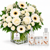 A large floral arrangement of white roses, white gerbera daisies, and green foliage in a clear square glass vase, displayed with three fragrance products from The Body Shop: a Santal Vanilla body mist, a jar, and a smaller bottle. A Flowrista tag hangs from the vase.