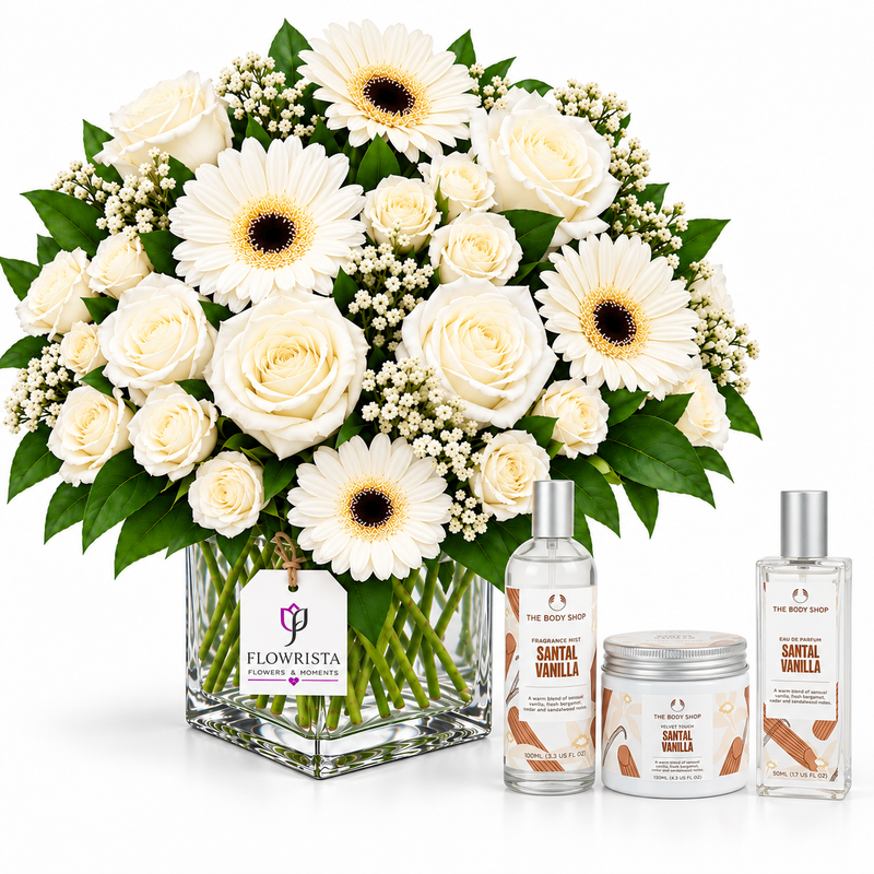A large floral arrangement of white roses, white gerbera daisies, and green foliage in a clear square glass vase, displayed with three fragrance products from The Body Shop: a Santal Vanilla body mist, a jar, and a smaller bottle. A Flowrista tag hangs from the vase.