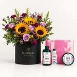 A curated gift set featuring a vibrant flower arrangement in a black round box labeled 'FLOWRISTA FLORAL ARTISANY' alongside The Body Shop bath and body items, including a pink gift box, a green bottle of British Rose body wash, a tube of hand cream, and a round tin of lip balm.