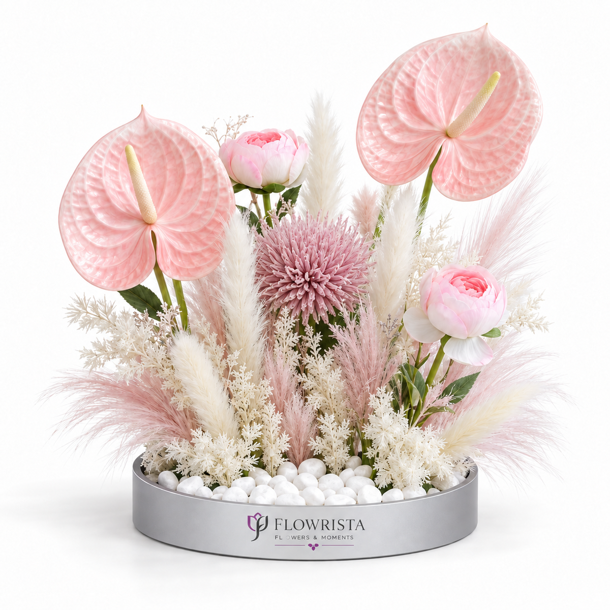 A floral arrangement in a round silver planter on a white background. The bouquet features light pink anthuriums, pink roses, a dusty pink chrysanthemum, and white and blush pampas grass.