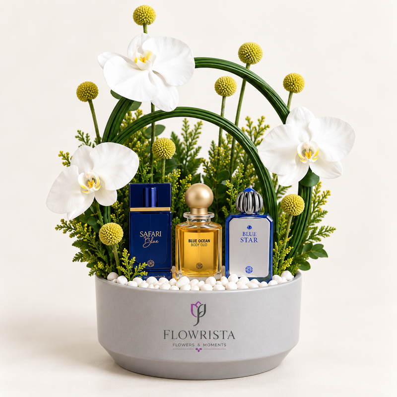 A floral gift arrangement in a round gray pot branded FLOWWRISTA, featuring white orchids, green foliage, yellow-green pom-pom flowers, and three perfume bottles: a dark blue Safari Blue bottle, a gold Blue Ocean bottle, and a blue Blue Star bottle, all arranged in white pebbles against a light beige background.