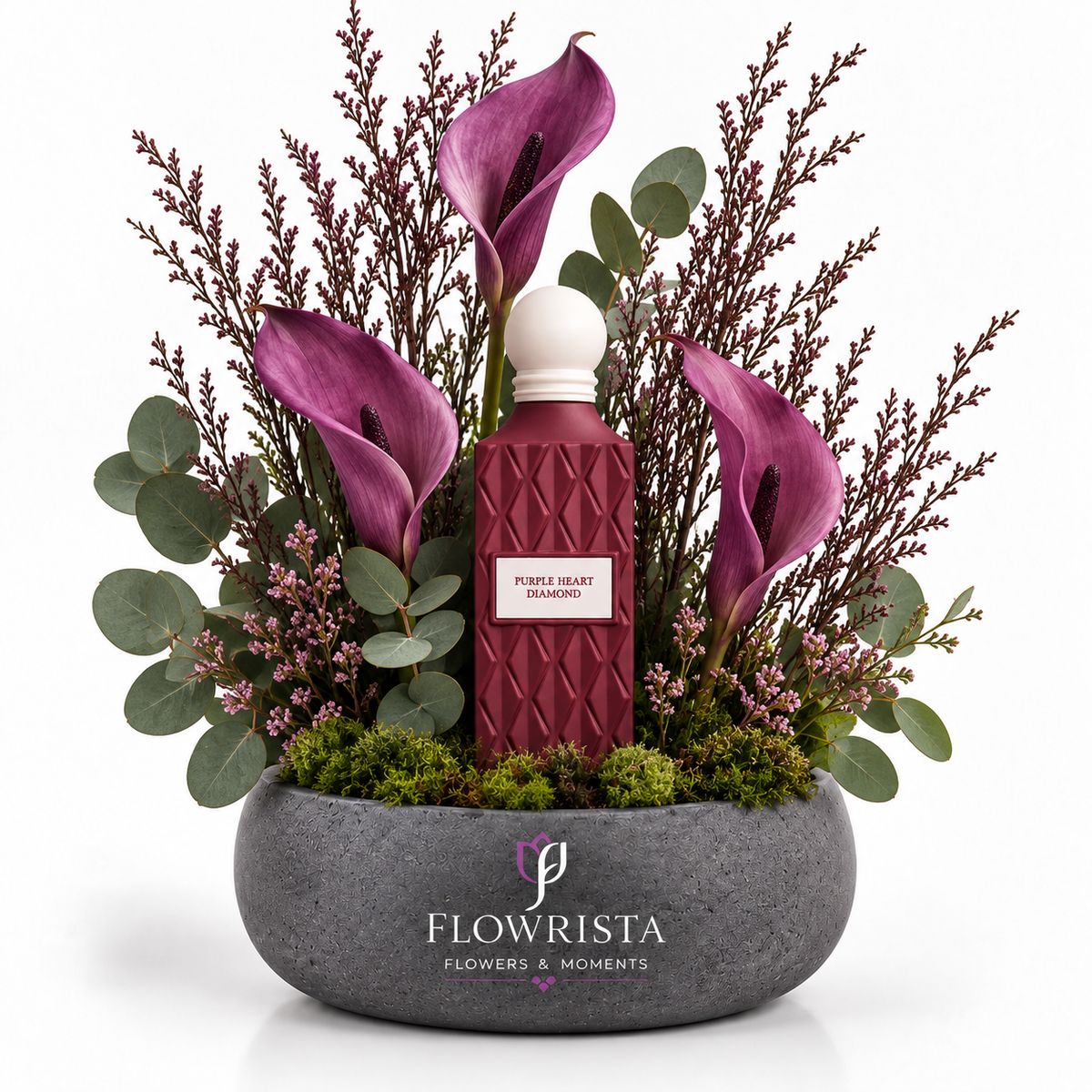 A floral arrangement in a round gray stone planter featuring a central textured burgundy bottle with a white cap and a label reading 'PURPLE BEAR ESSENTIALS,' surrounded by purple calla lilies, green eucalyptus, and pinkish filler stems on a white background. The planter is branded FLOWRISTA FLOWERS & MOMENTS.