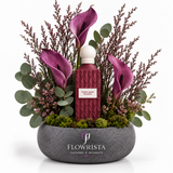 A floral arrangement in a round gray stone planter featuring a central textured burgundy bottle with a white cap and a label reading 'PURPLE BEAR ESSENTIALS,' surrounded by purple calla lilies, green eucalyptus, and pinkish filler stems on a white background. The planter is branded FLOWRISTA FLOWERS & MOMENTS.
