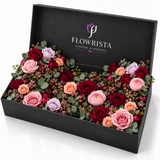 A black floral gift box from Flowrista Flowers & Moments, open to reveal a dense arrangement of pink, burgundy, peach, and lavender roses with green eucalyptus leaves, photographed on a white background.