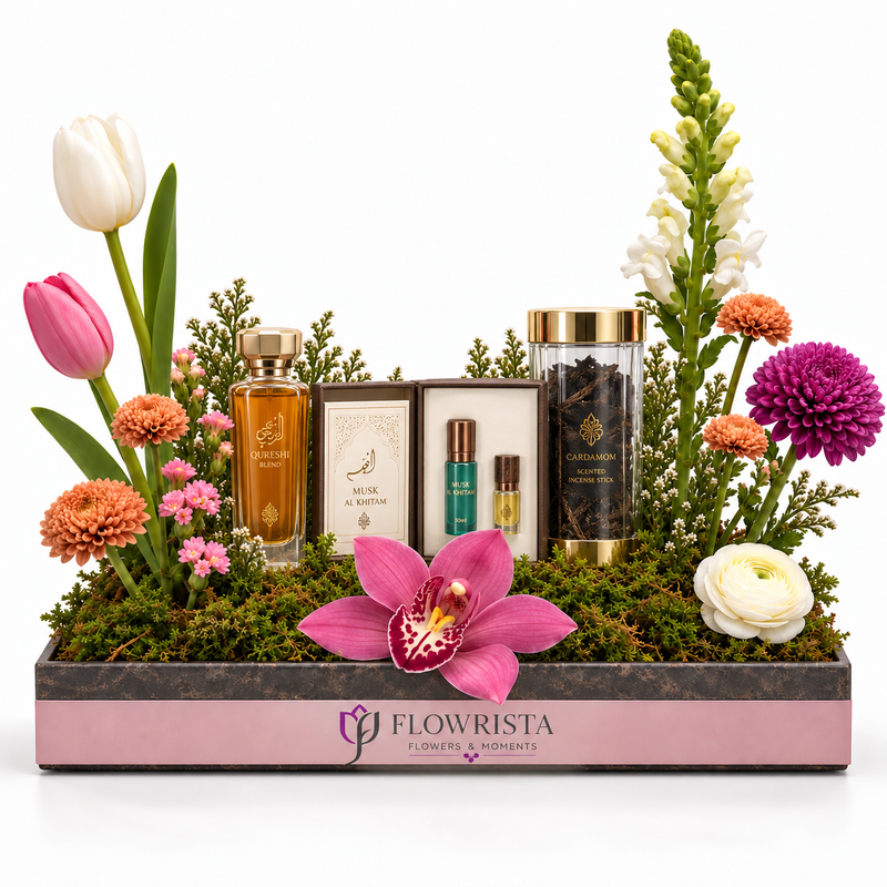 A floral gift basket arrangement by Flowrista featuring various perfume bottles, a candle, and a jar of essential oils nestled in green moss and surrounded by colorful flowers, including tulips, an orchid, and chrysanthemums, in a pink and dark gray wooden tray.