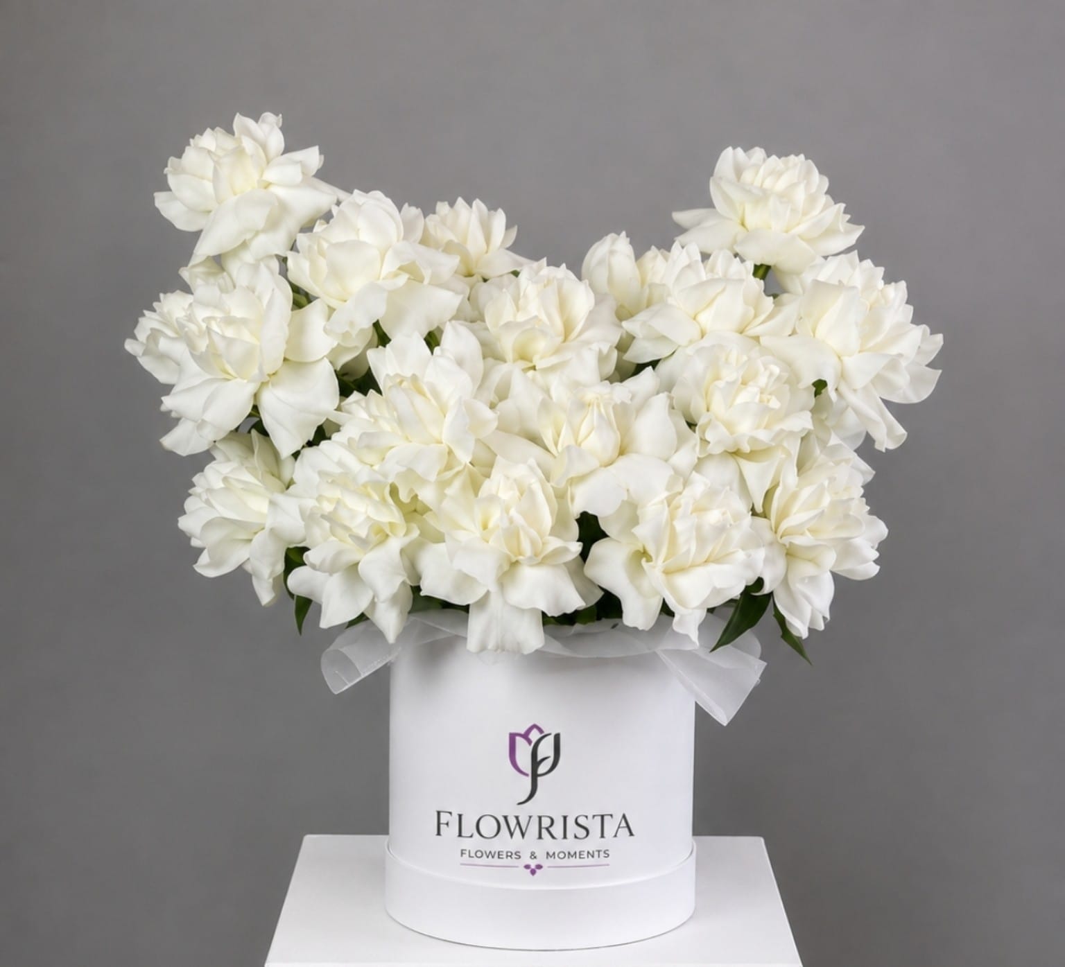 A large bouquet of white carnations arranged in a round white box with a ribbon and the visible brand name FLOWWRISTA, photographed against a plain gray studio background.