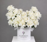 A large bouquet of white carnations arranged in a round white box with a ribbon and the visible brand name FLOWWRISTA, photographed against a plain gray studio background.