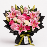 A vibrant flower bouquet featuring pink lilies, pink and white roses, and pink hydrangeas wrapped in black paper with a green ribbon and a 'FLOWWISTA' tag, set against a plain white background.