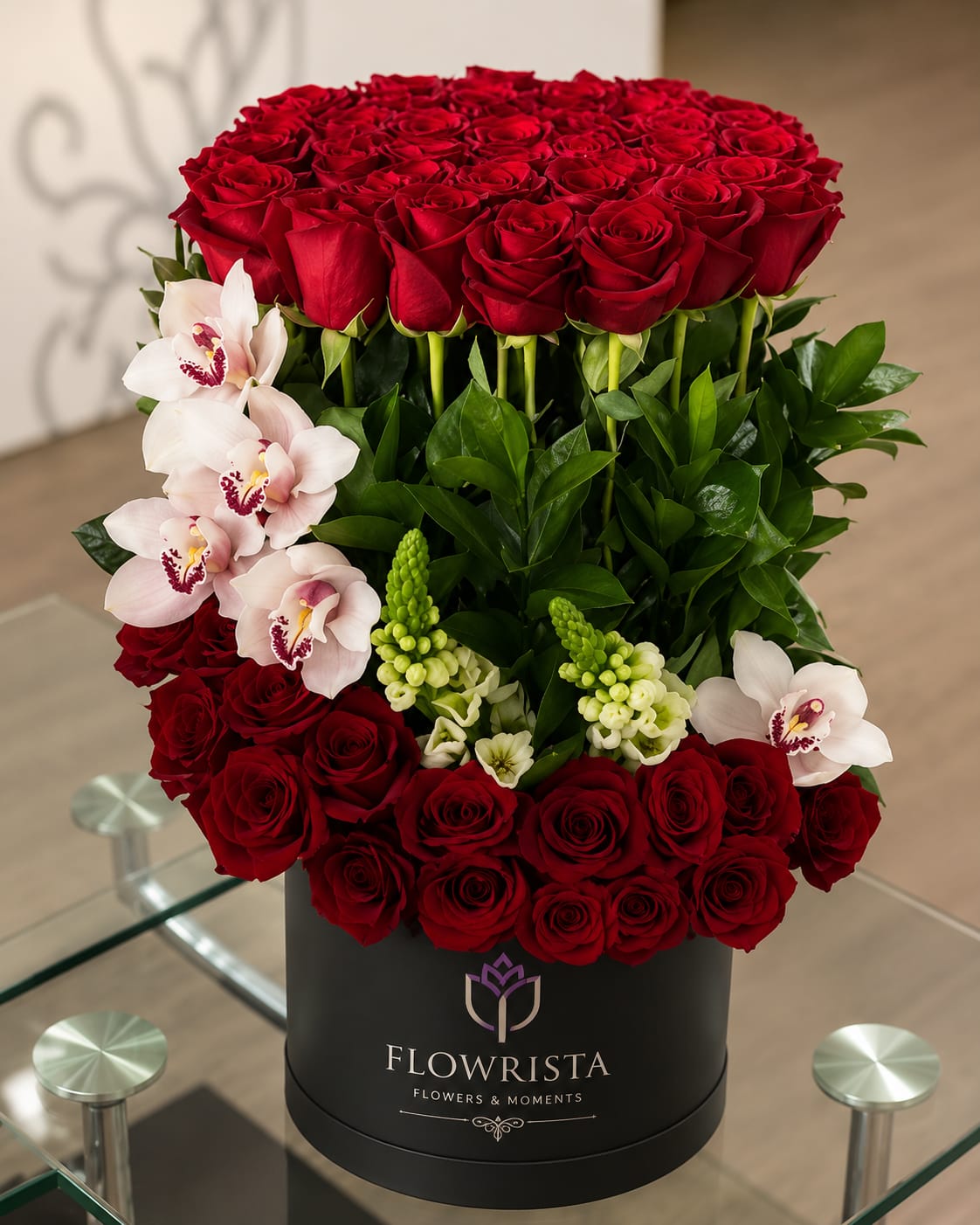 A large floral arrangement of red roses, white orchids, and green foliage in a black cylindrical container branded FLOWRISTA, placed on a glass table in an indoor setting.