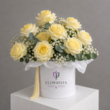 A round white gift box filled with pale yellow roses, green eucalyptus leaves, and small white baby's breath flowers, with a tassel and the visible brand name FLOWWRISTA on the front, displayed on a white pedestal against a gray background.