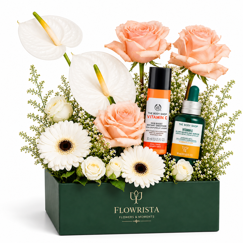 A floral gift box arrangement from Flowrista featuring white and peach roses, white anthuriums, and white gerbera daisies in a dark green box, styled with two skincare products: a The Body Shop Vitamin C serum and a The Body Shop Rosehip treatment.