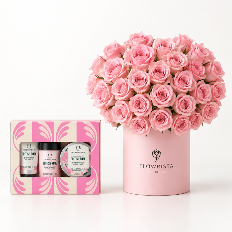 A pink floral gift set featuring a large bouquet of pink roses in a pink cylindrical box labeled FLOWWRISTA, alongside a pink and white patterned gift box containing two small bottles and a jar from The Body Shop British Rose.