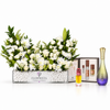 A floral arrangement in a rectangular glass planter with white flowers and green foliage, displayed alongside perfume bottles and a branded Flowerista box on a clean white background.