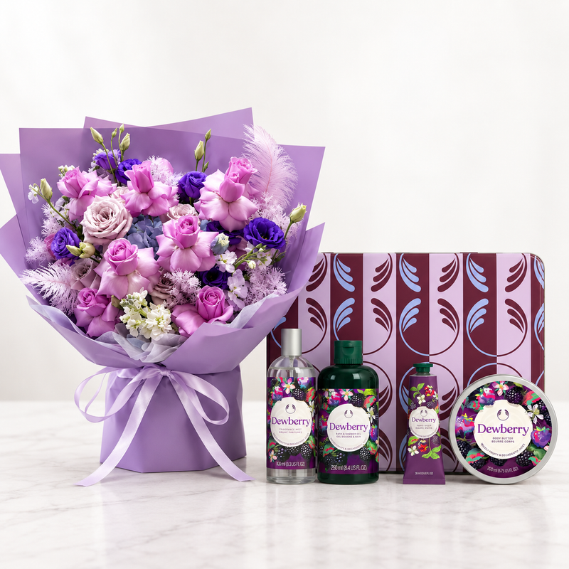 A luxury gift set featuring a purple and pink flower bouquet wrapped in lavender paper, alongside bottles and a tin of Dewberry bath and body products on a white marble surface against a white background.