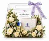A clear acrylic gift basket from Flowrista, decorated with white roses, baby's breath, and green eucalyptus leaves, tied with a lavender ribbon. Inside are a small amber perfume bottle with a blue cap, a white patterned box, and a floral arrangement.