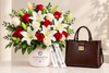 A floral arrangement in a white Flowerista vase with red and white flowers, green foliage, and two white Flowerista ribbons, placed beside a dark brown textured handbag on a light marble surface against a warm beige wall.
