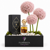 A luxury fragrance gift set by Flowrista, featuring a bottle of Cardamom Body Oud Eau de Parfum with amber liquid and a gold cap, displayed next to its black textured box. The set is beautifully arranged in a black rectangular box filled with white stones, green moss, and three pink allium flowers, set against a clean white background.