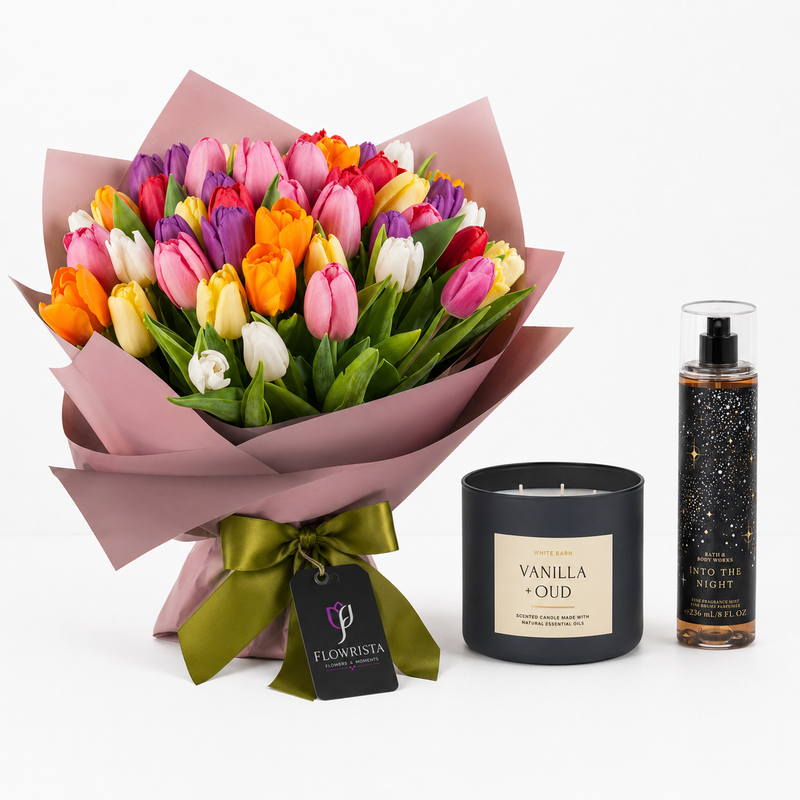 A gift set featuring a vibrant bouquet of multicolored tulips in pink, yellow, orange, and purple, wrapped in dusty pink paper with a green ribbon and a 'Flowrista' tag. Next to it are a black jar candle labeled 'Vanilla + Oud' and a clear bottle of 'Into the Night' body spray on a clean white background.