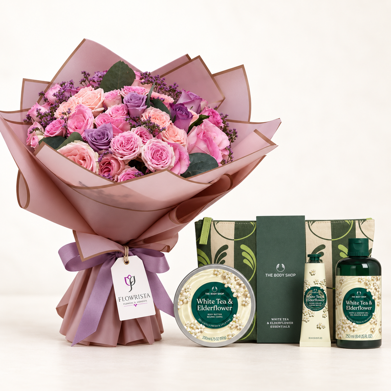 A floral gift set featuring a large bouquet of pink and purple roses wrapped in dusty pink paper, accompanied by a round tin of White Tea & Elderflower bath bomb, a green box of The Body Shop, a hand cream bottle, and a white beach deodorizer bottle on a clean white background.