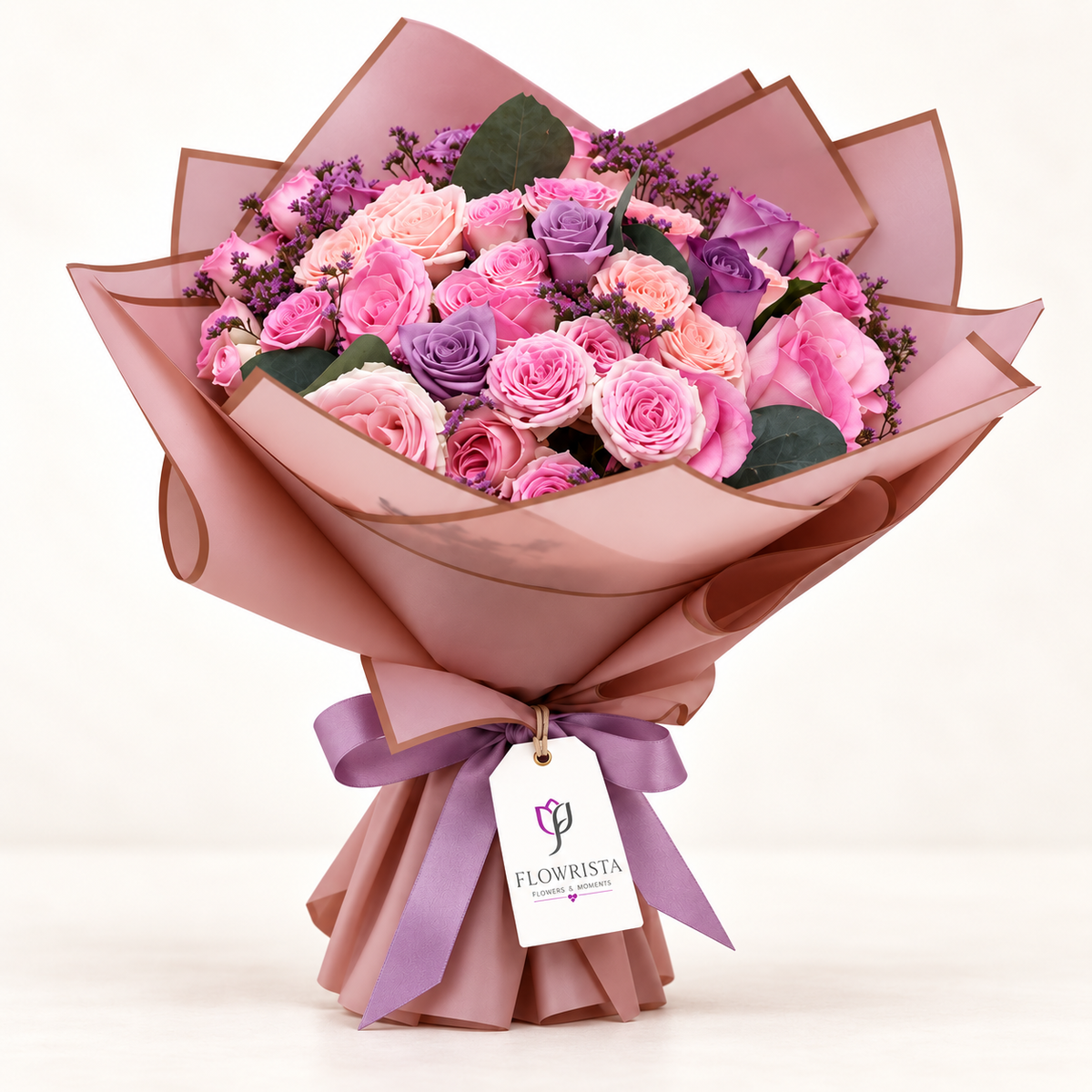 A vibrant bouquet of pink and purple roses wrapped in dusty pink paper with a purple ribbon and a 'FLOWRISTA' tag, set against a clean white background.