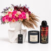 A curated gift set featuring a vibrant pink and red floral arrangement in a white female bust vase, a black candle labeled Vanilla Oud, a black body spray can labeled Adrenaline, and a black tag from Flowrista.