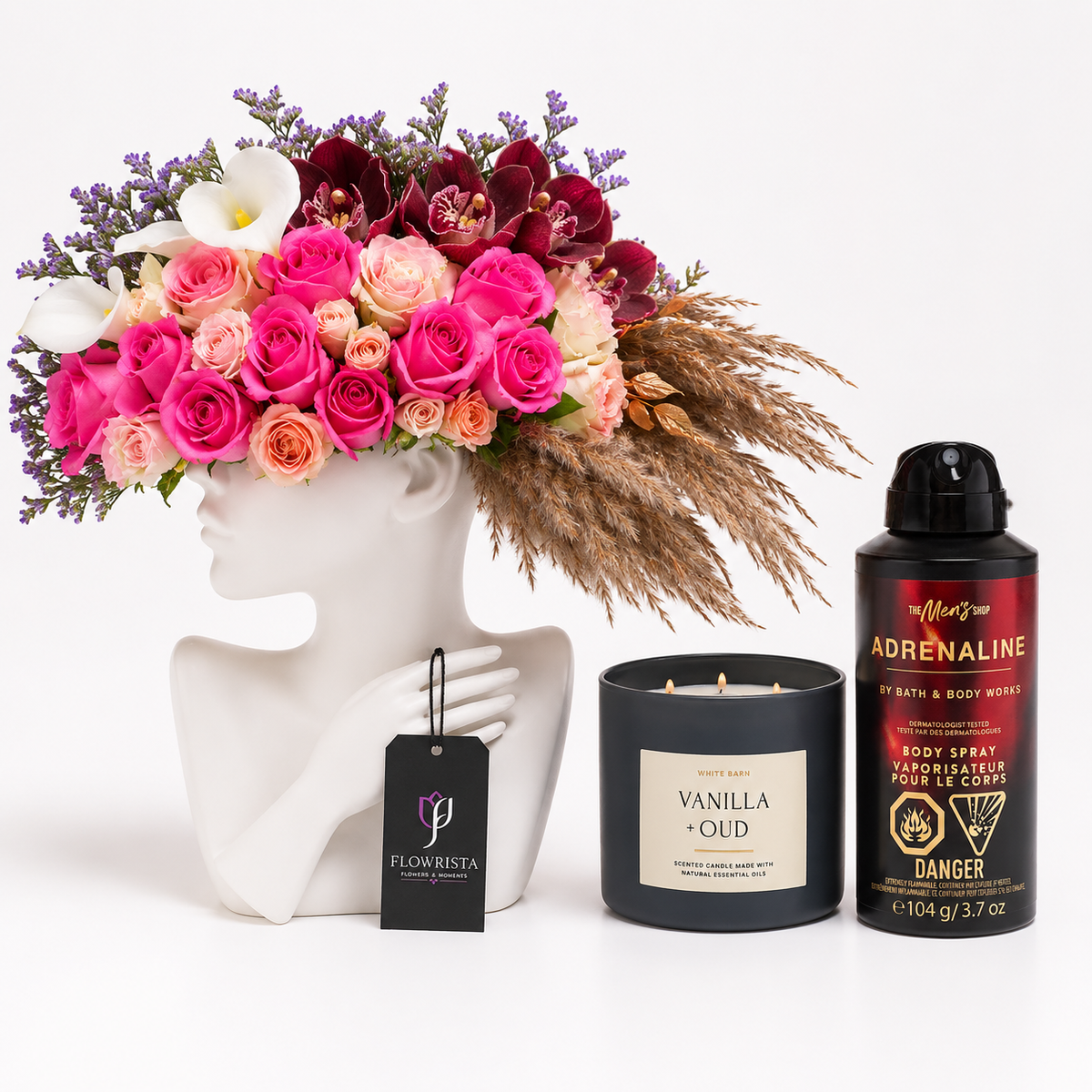 A curated gift set featuring a vibrant pink and red floral arrangement in a white female bust vase, a black candle labeled Vanilla Oud, a black body spray can labeled Adrenaline, and a black tag from Flowrista.
