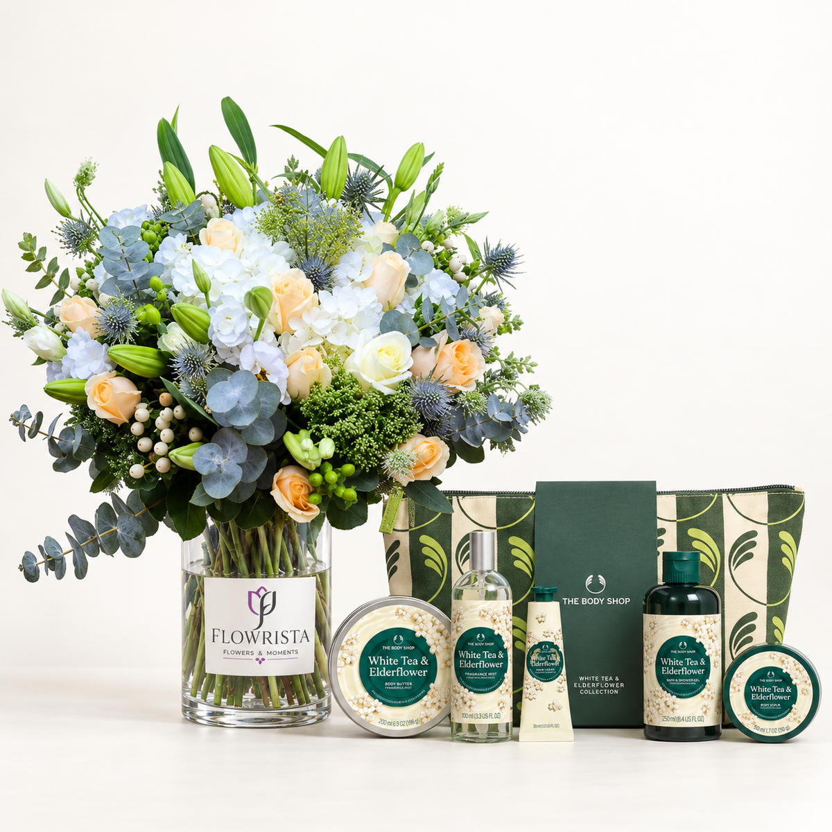 A curated gift set featuring a large floral arrangement in a glass vase labeled FLOWWRISTA, alongside The Body Shop bath and body products including a white tea & elderflower shower foam bottle, hand cream tube, body wash bottle, and body butter tin, arranged on a white surface against a clean white background.